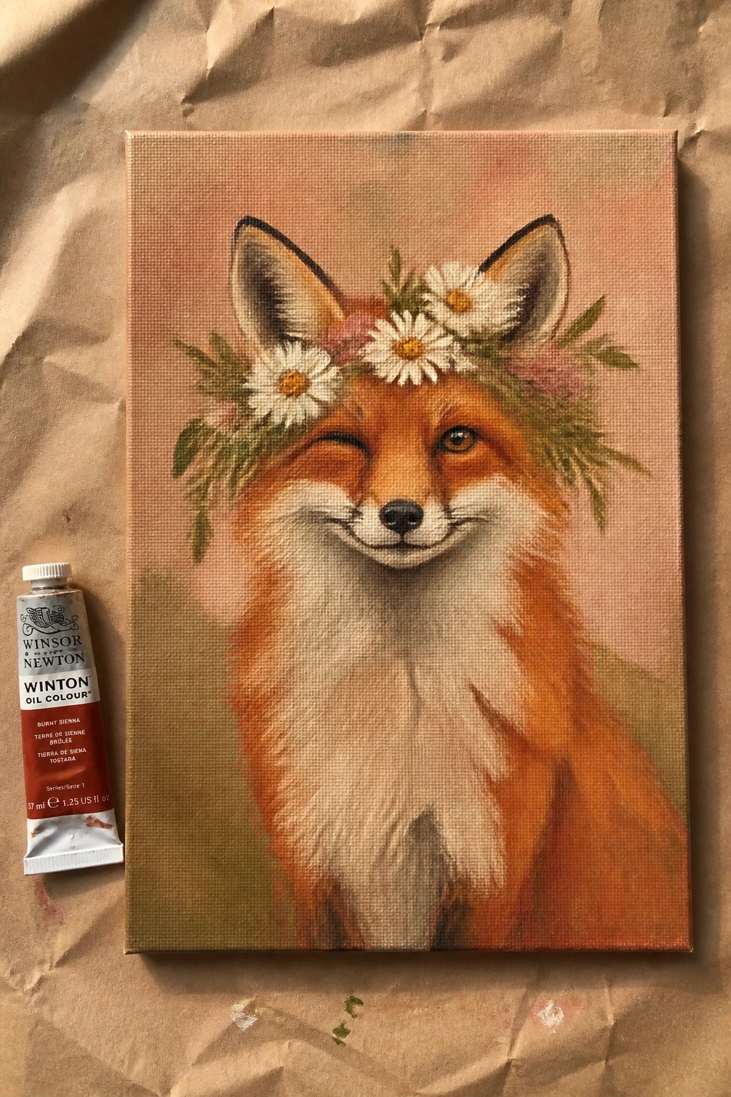 Painting of winking fox with daisy flower crown beside oil paint tube on paper