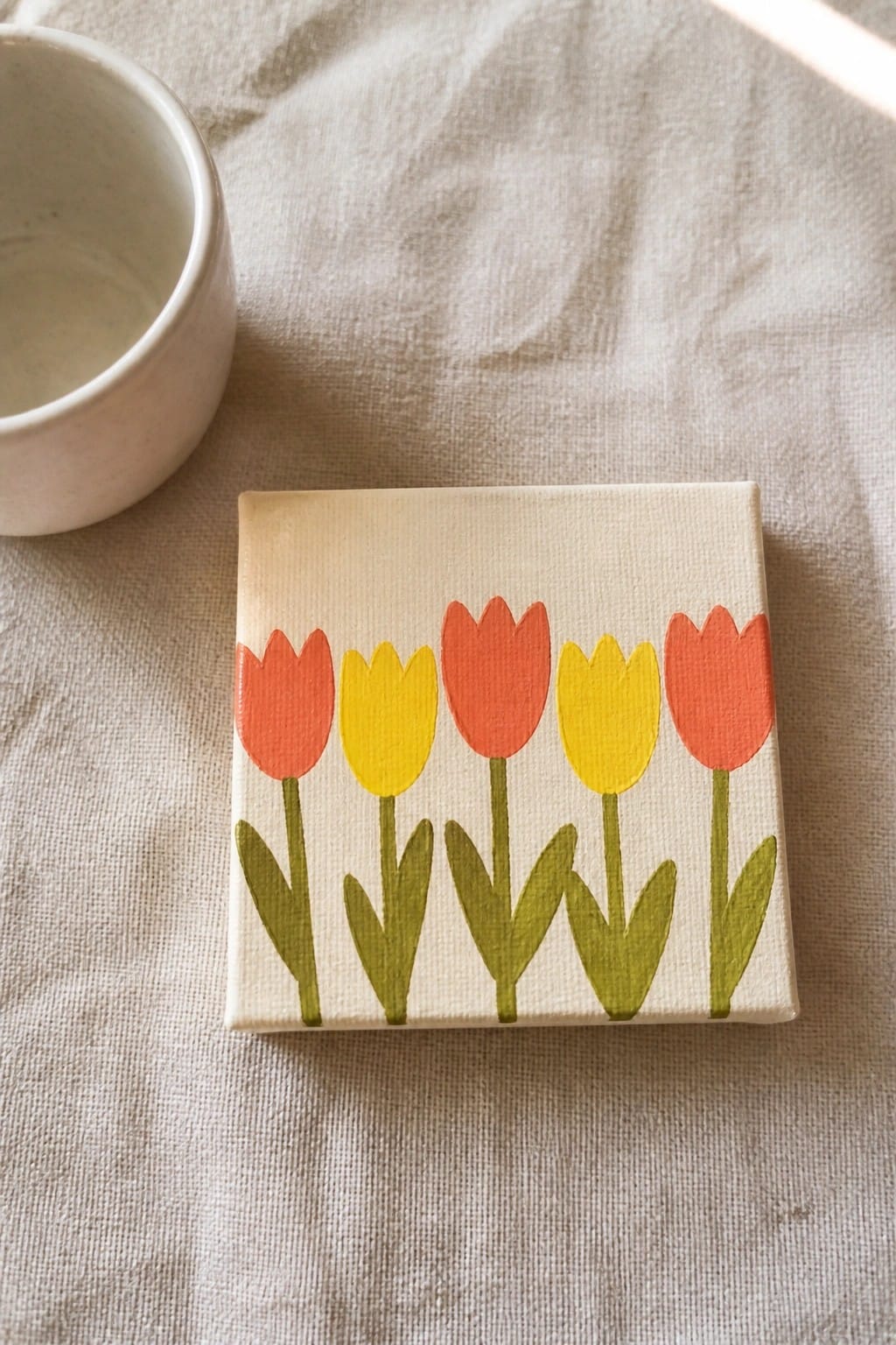 Small canvas with five red and yellow tulips painted on beige fabric beside white bowl.