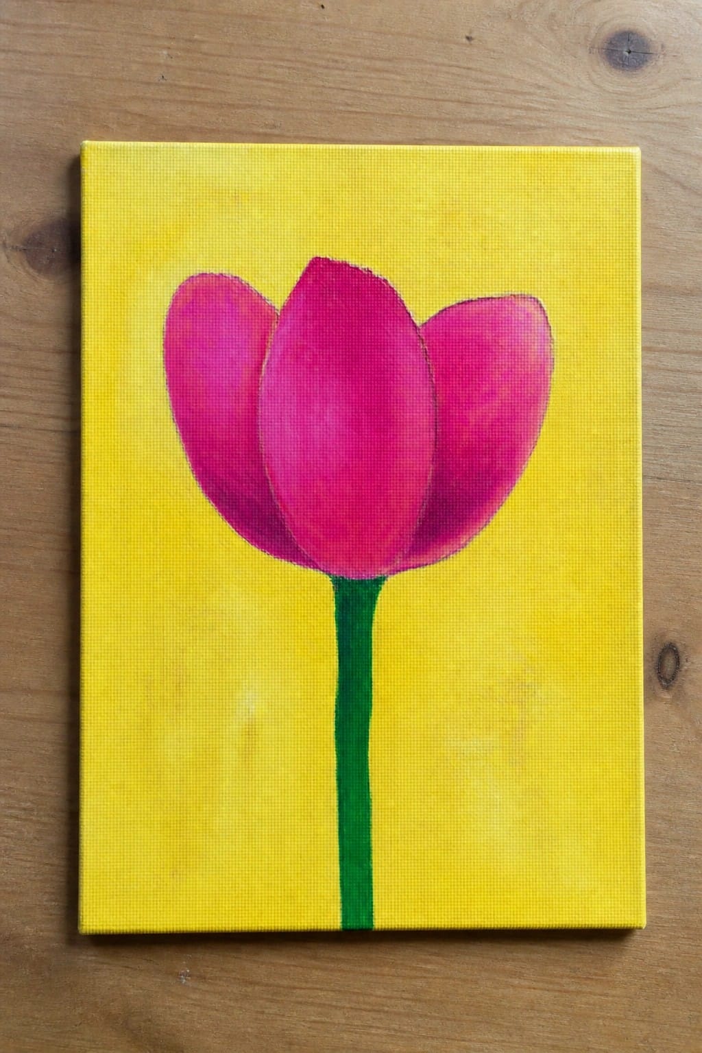 Pink tulip with green stem on yellow canvas atop wooden surface
