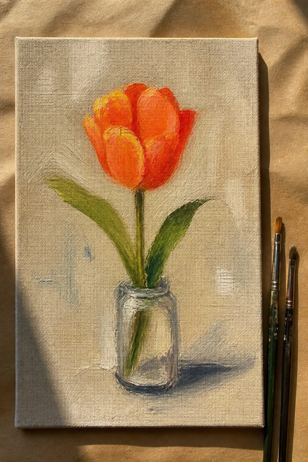 Orange tulip in glass jar painted on canvas beside brushes