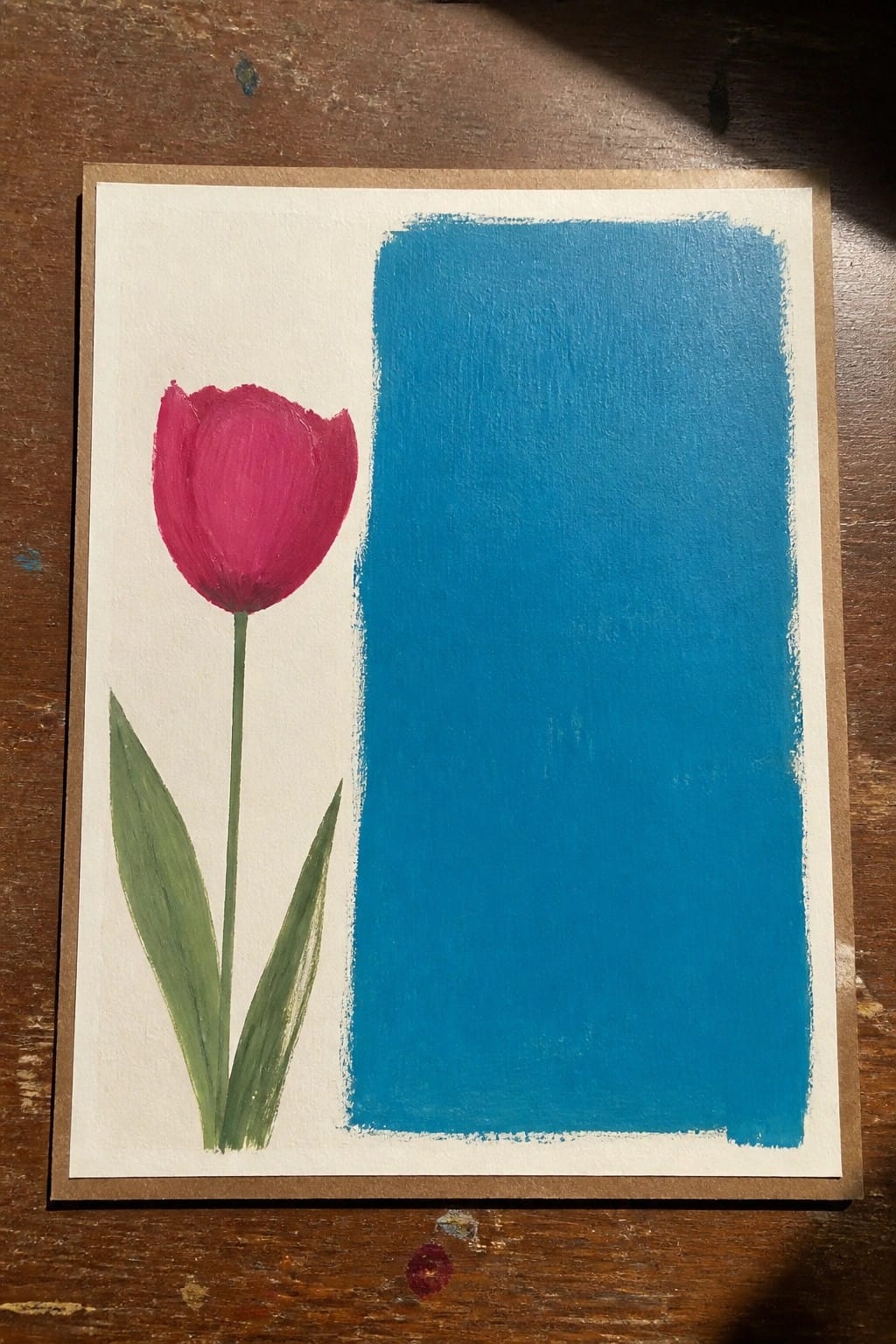 Pink tulip with green stem and leaves beside blue rectangle on white paper