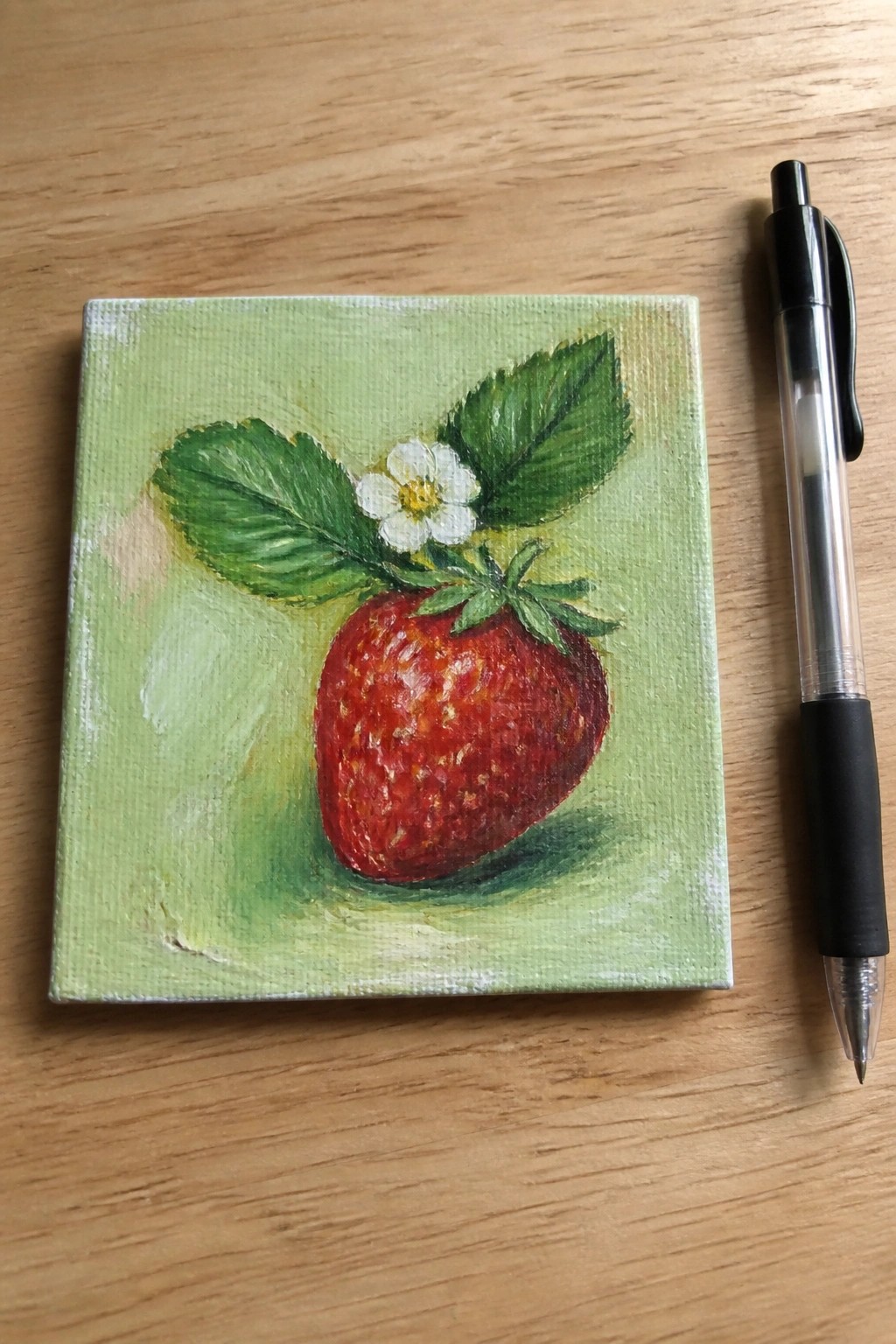 Red strawberry with leaves and flower on small green canvas beside pen on wood.