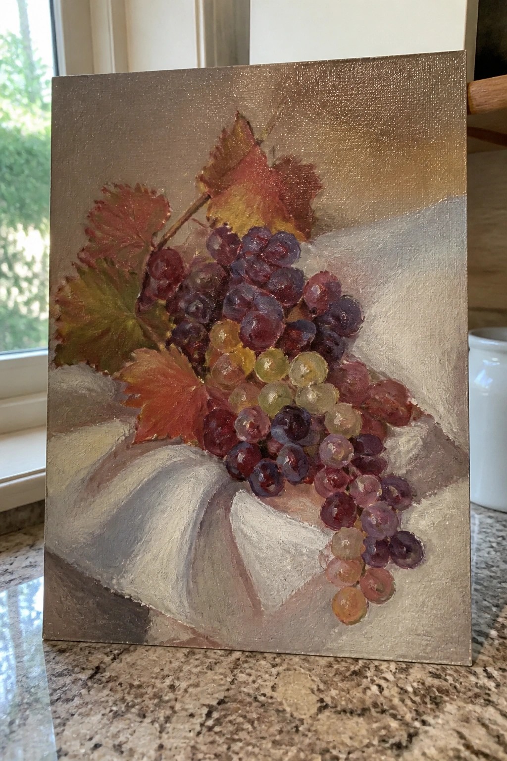 Still life painting of multicolored grapes and autumn leaves on white cloth.