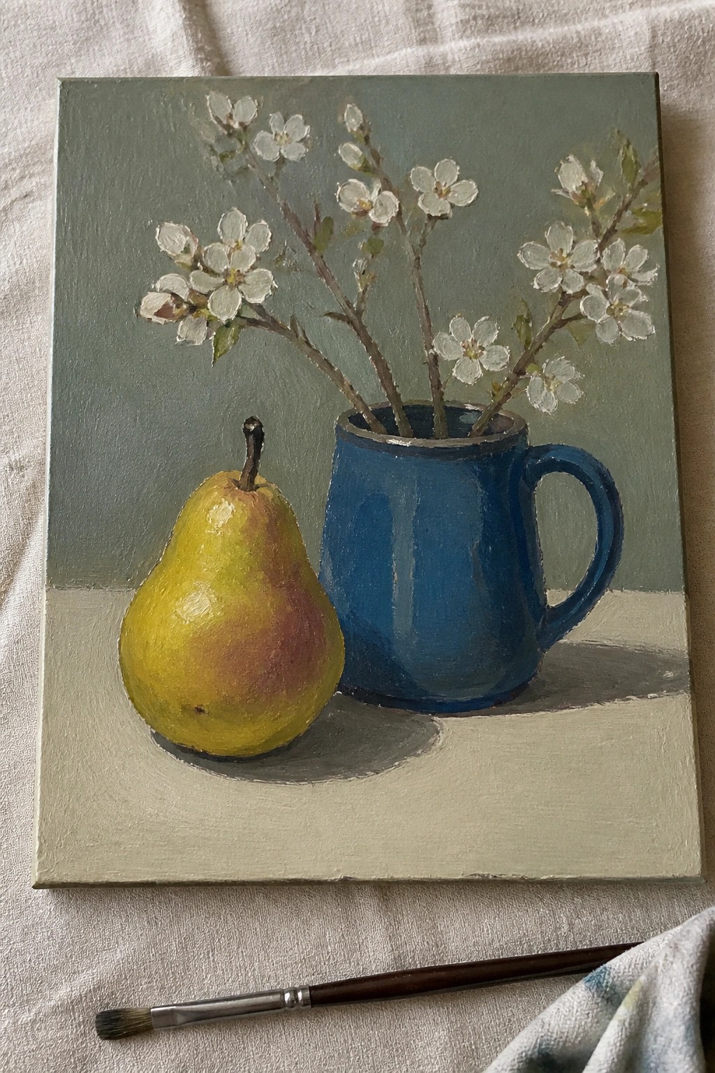 An oil painting of a yellow pear beside a blue mug holding white flowering branches on a light surface with a muted green background.