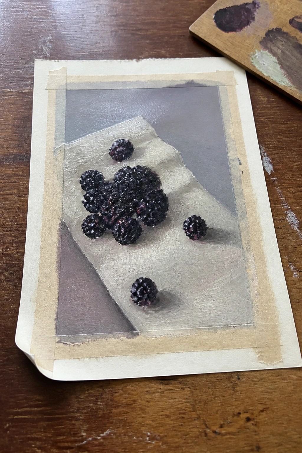 Realistic painting of blackberries on white paper taped to wooden table with palette nearby