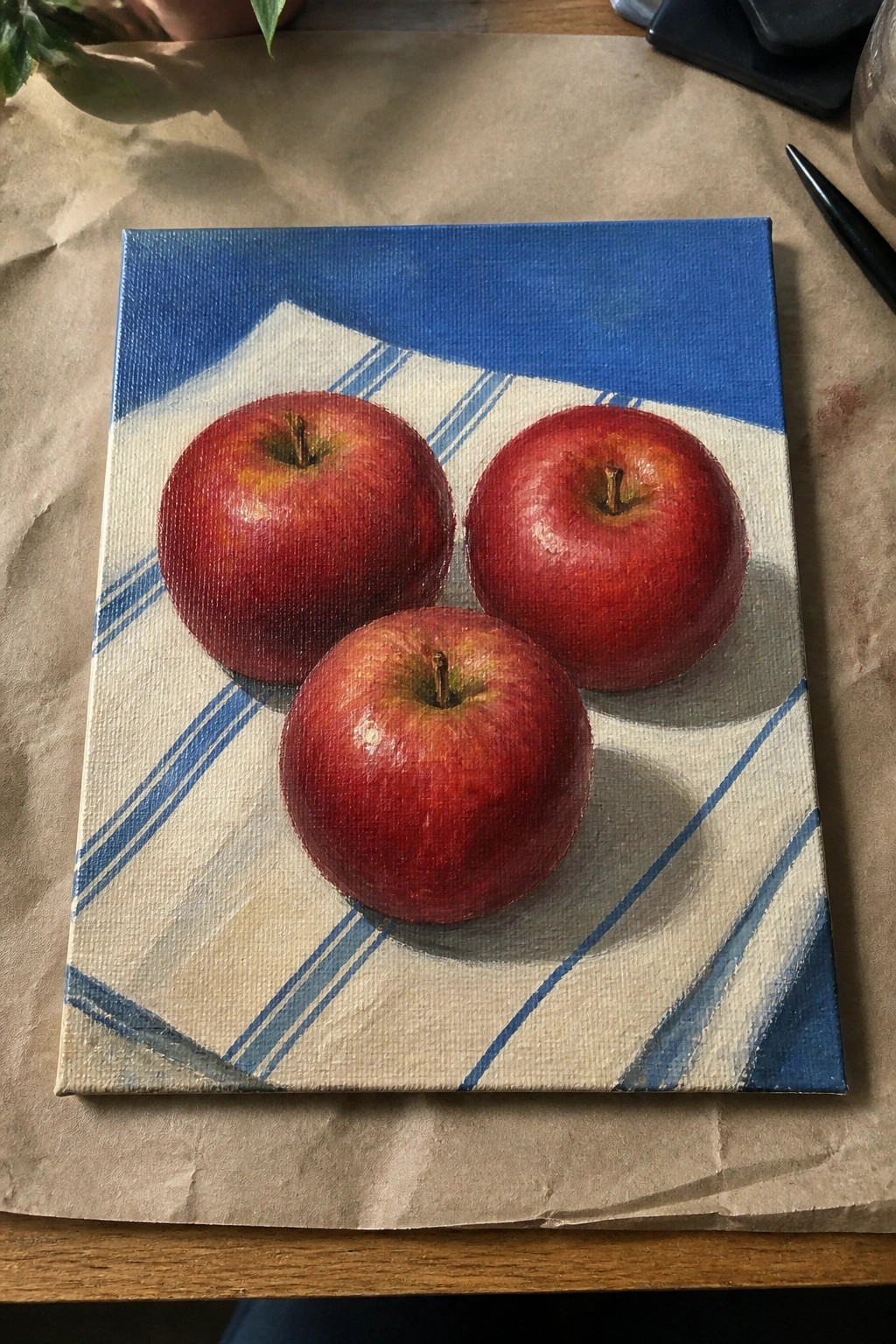 Three glossy red apples on blue-striped cloth atop a canvas painting
