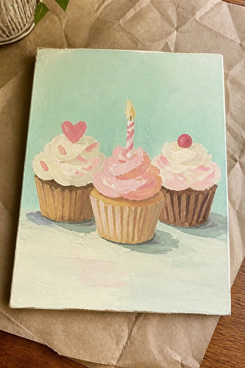 Three painted cupcakes with pink frosting, heart, cherry, and lit candle on mint canvas.