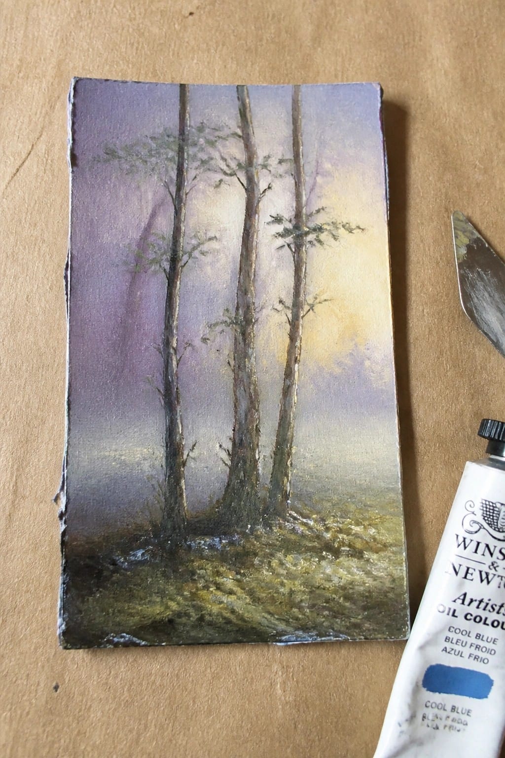 Miniature misty forest painting beside Winsor & Newton cool blue oil paint tube and knife