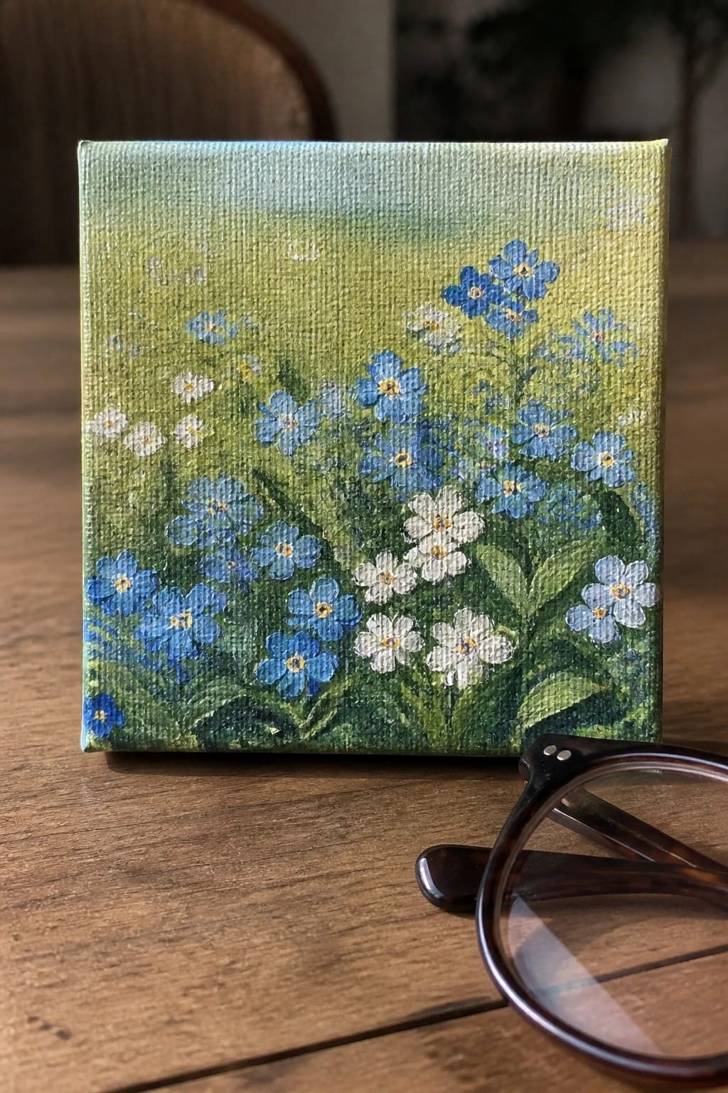 Small canvas painting of blue and white flowers with glasses on wooden table.