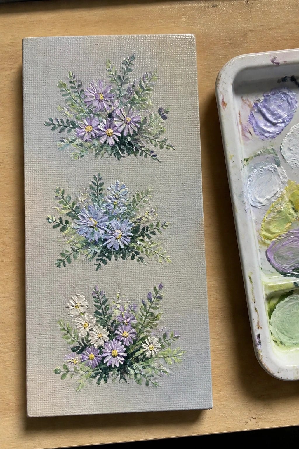 Three stacked floral bouquets in purple, blue, and white on canvas beside palette