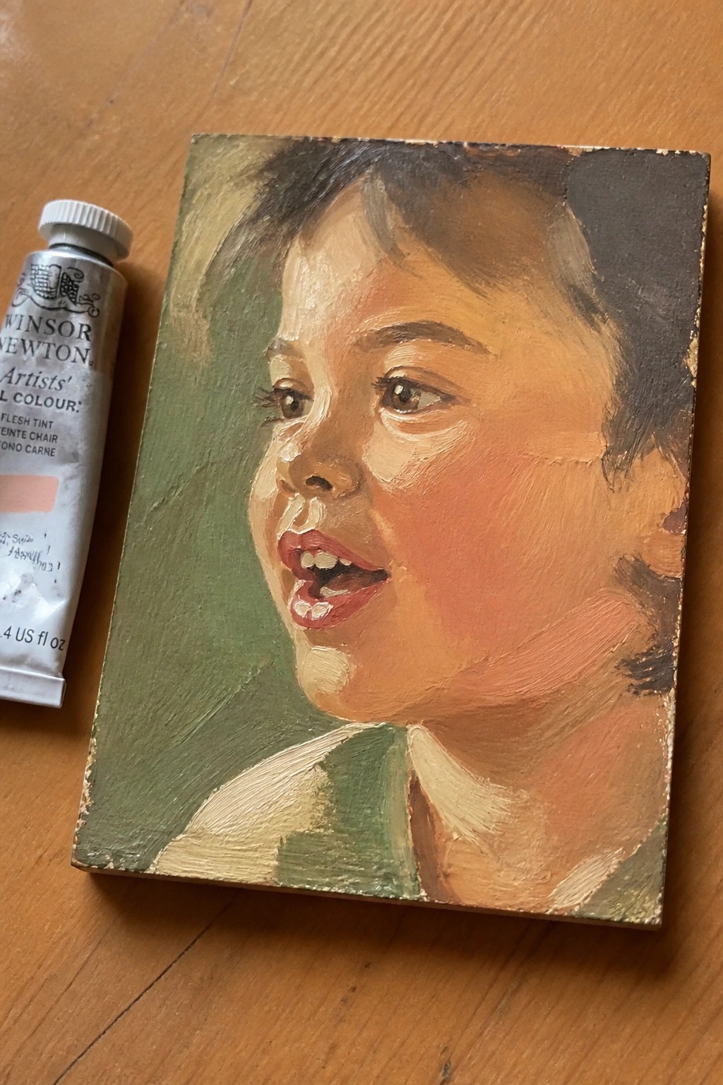 Oil portrait of child's face beside Winsor & Newton flesh tint paint tube on wood.