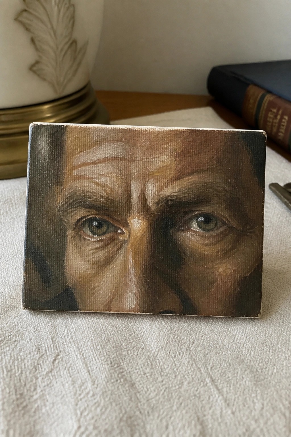 Small canvas painting of older man's eyes beside blue book and white vase