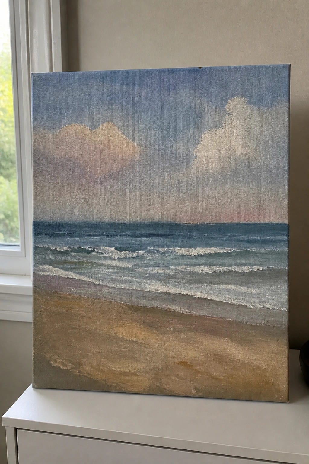 Painting of sandy beach, blue ocean waves, and cloudy sky on canvas by window.