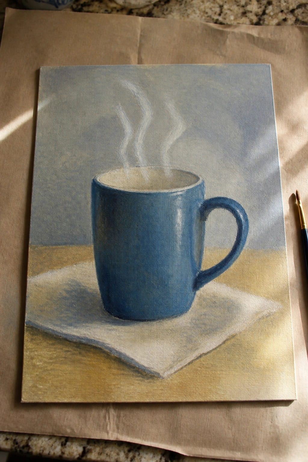 Steaming blue mug on white napkin atop wooden surface, paintbrush beside canvas painting.