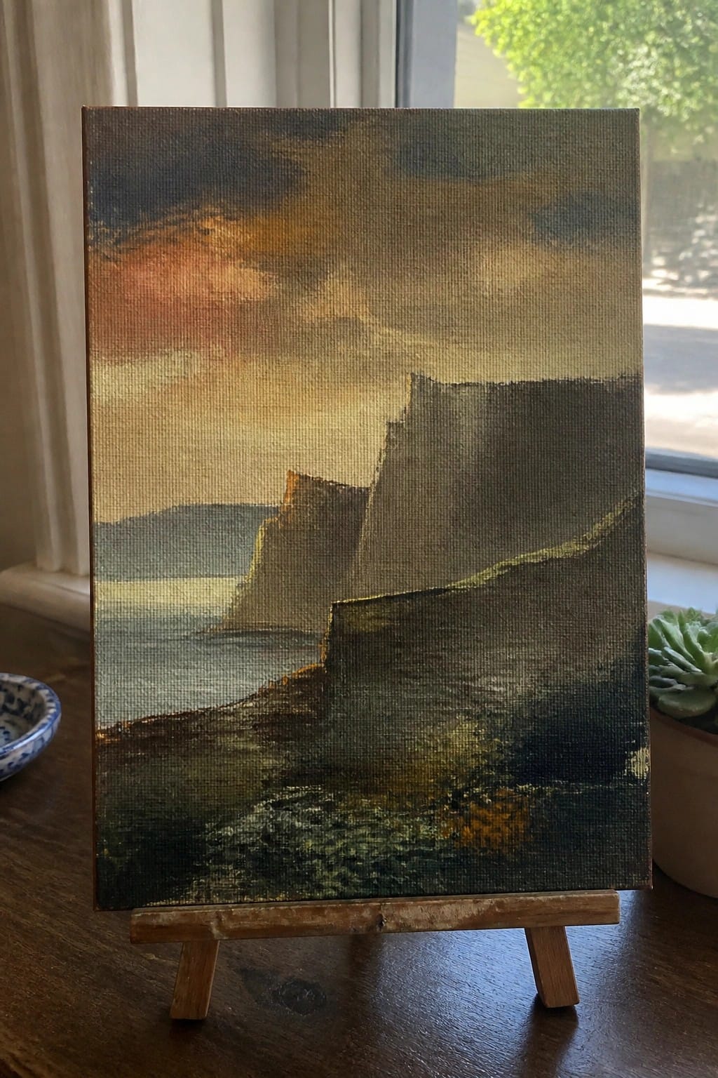 An oil painting on a small canvas shows abstract cliffs and a colorful sky with blended oranges and darks, resting on a wooden easel near a window.