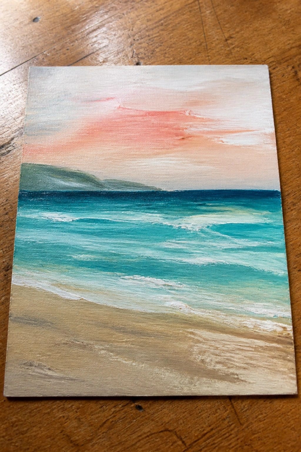 Painted seascape of turquoise waves on sandy beach under pink-orange sunset sky with hills.