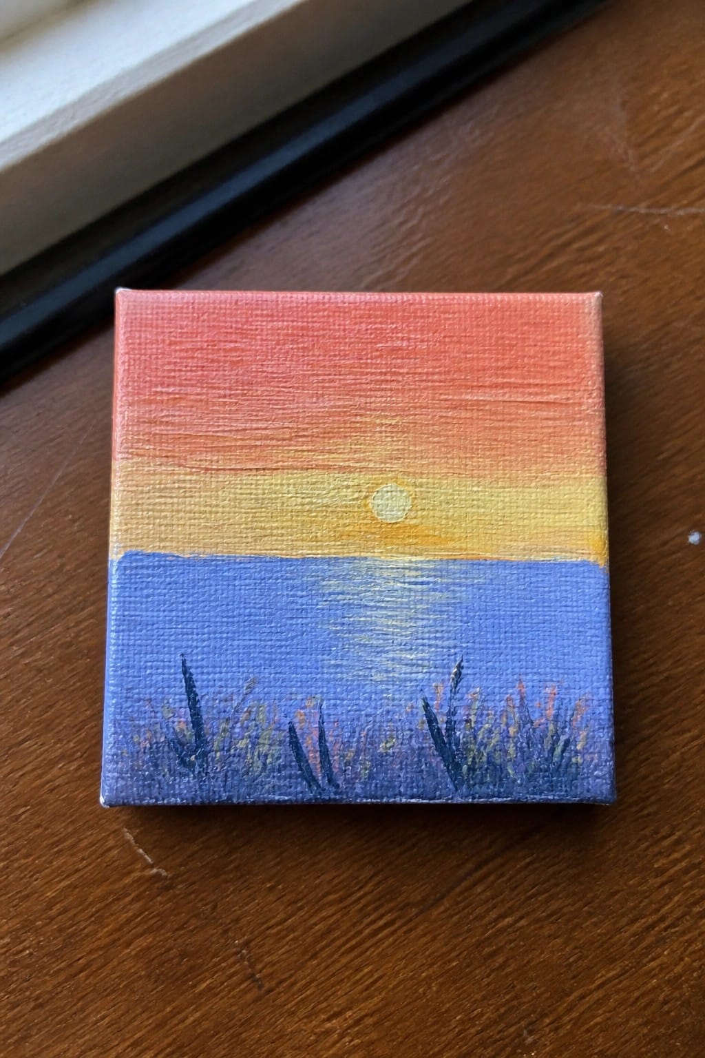 A small square oil painting showing a sunset gradient sky over blue water with a yellow sun and dark grass strokes at the bottom.