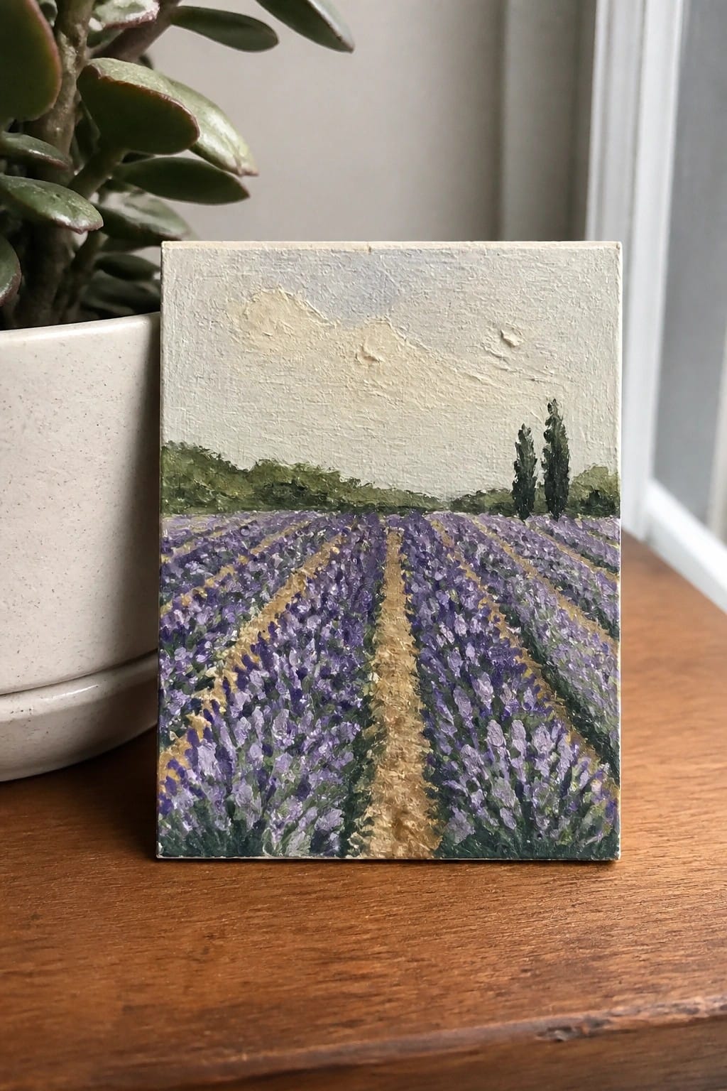Small lavender field painting with purple rows beside potted plant on wood table