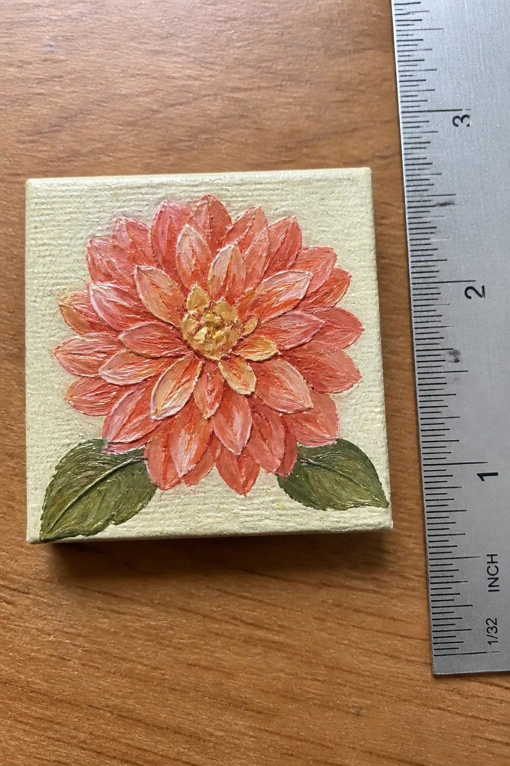 Miniature canvas with orange-red dahlia flower, yellow center, and green leaves beside ruler