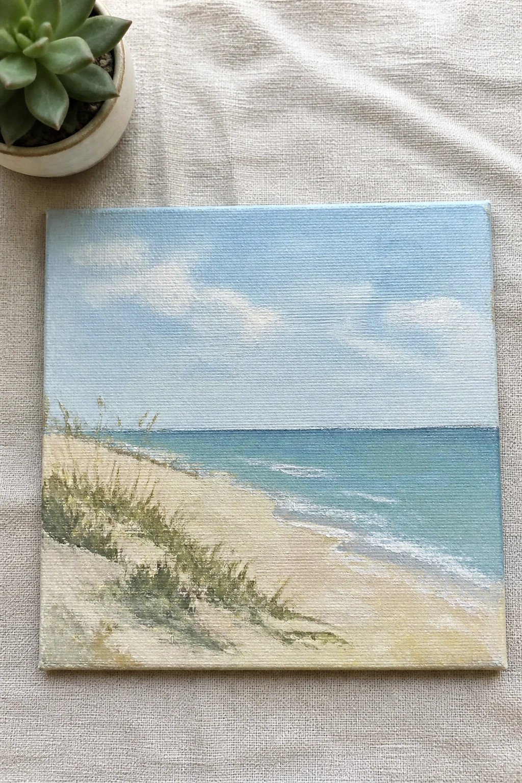 Square canvas beach painting with blue sky and turquoise sea beside potted succulent