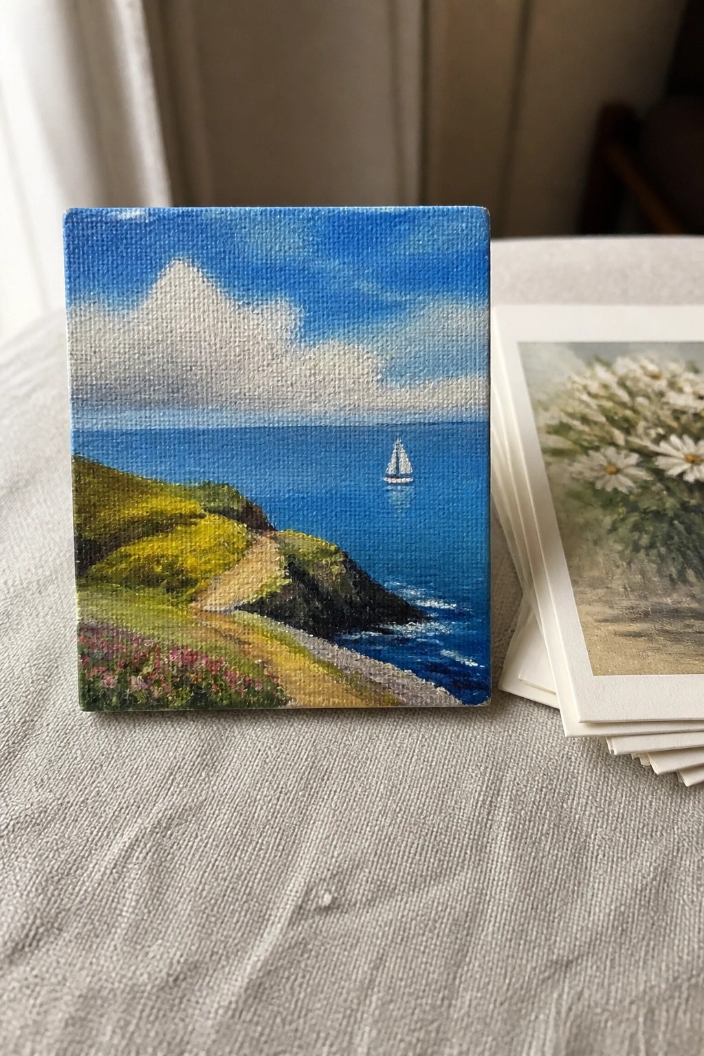 Small canvas painting of blue ocean with sailboat beside stack of daisy cards
