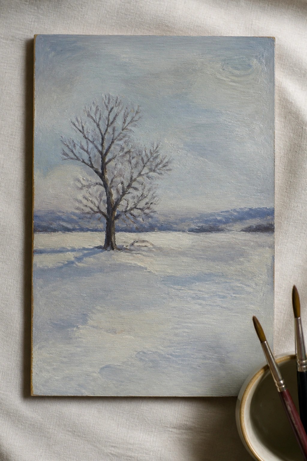 Winter landscape painting of bare tree in snowy field with paintbrushes nearby