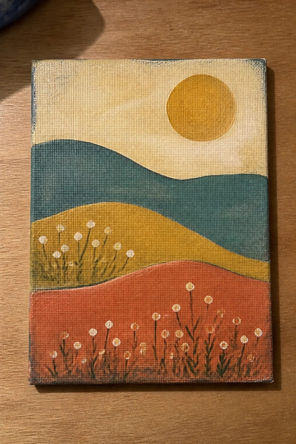Abstract painting of yellow sun over wavy teal, yellow, and orange hills with white flowers on wood.