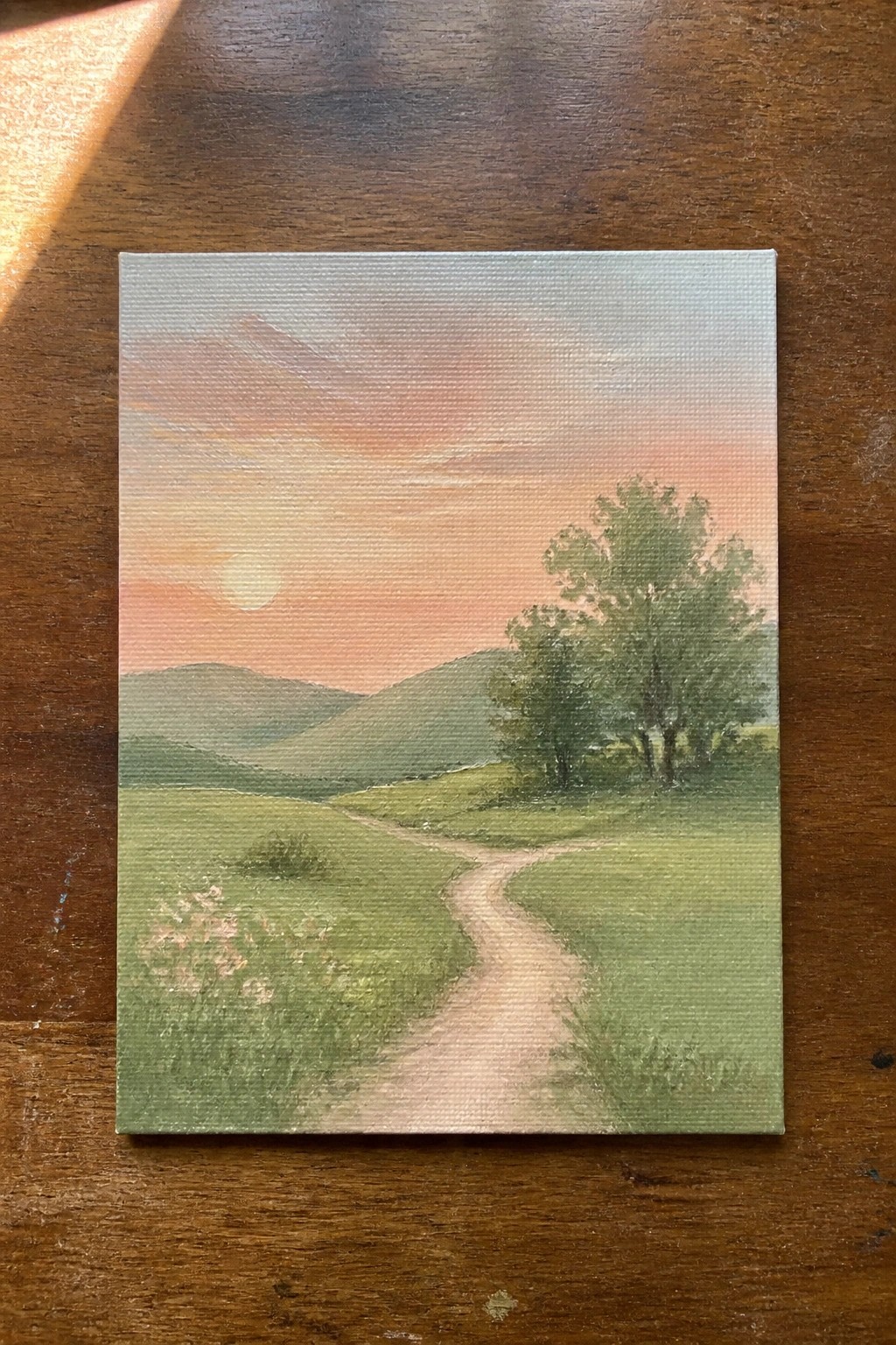 Winding dirt path curves through green meadows toward hills at pink-orange sunset.