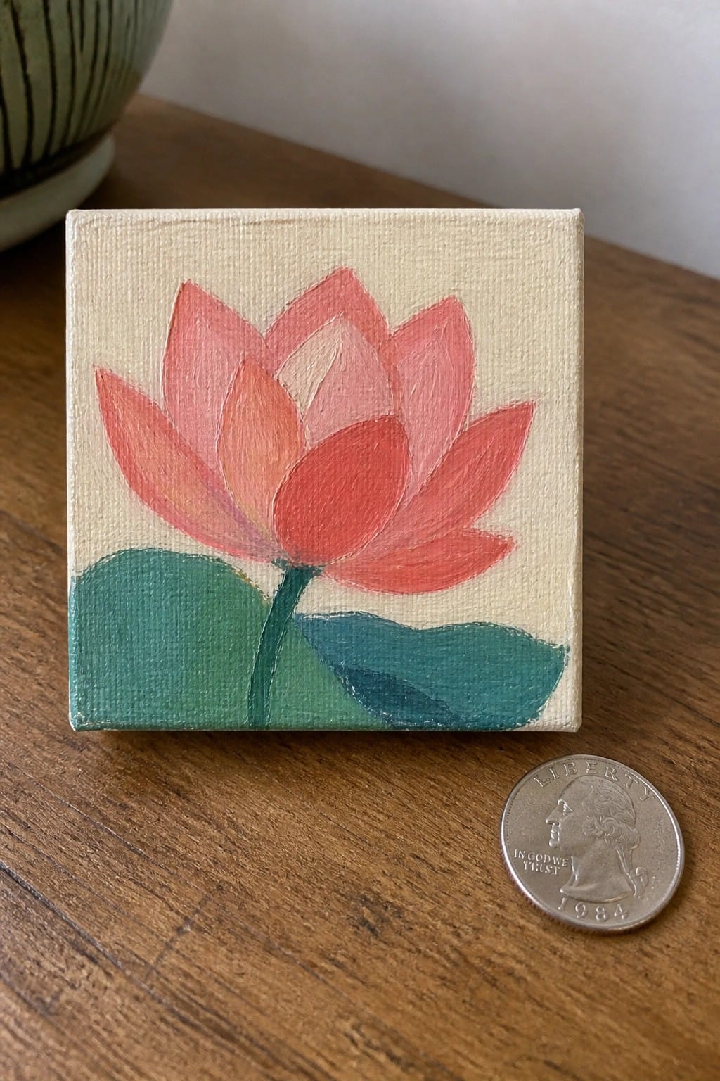 Small pink lotus painting on canvas beside 1984 quarter on wooden table