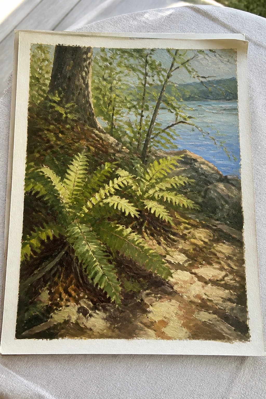 Oil painting of ferns by tree trunk on rocky shore overlooking blue lake and hills.