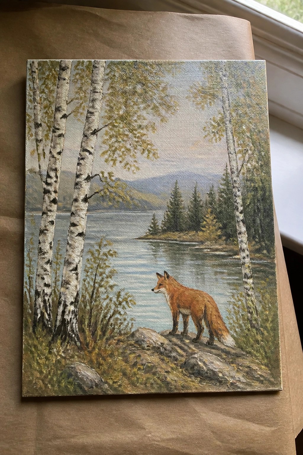 Red fox stands on rocks by calm lake with birch trees and distant mountains.