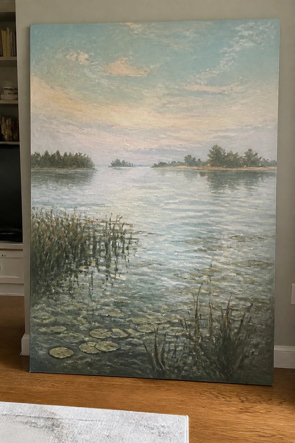 Large oil painting of serene lake with lily pads, reeds, and distant islands under pastel sky.