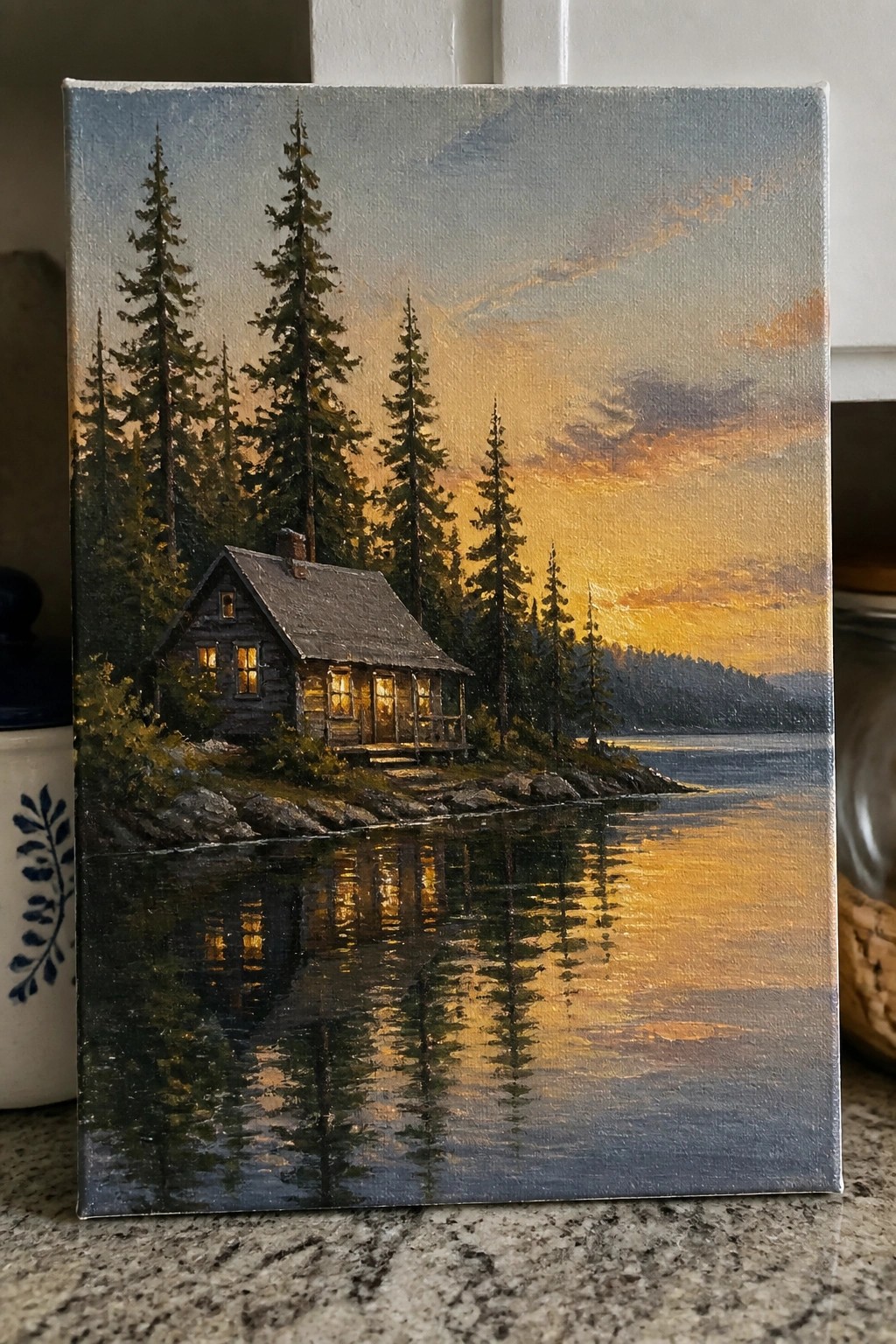 Wooden cabin with lit windows reflected in calm lake at golden sunset amid tall pines.