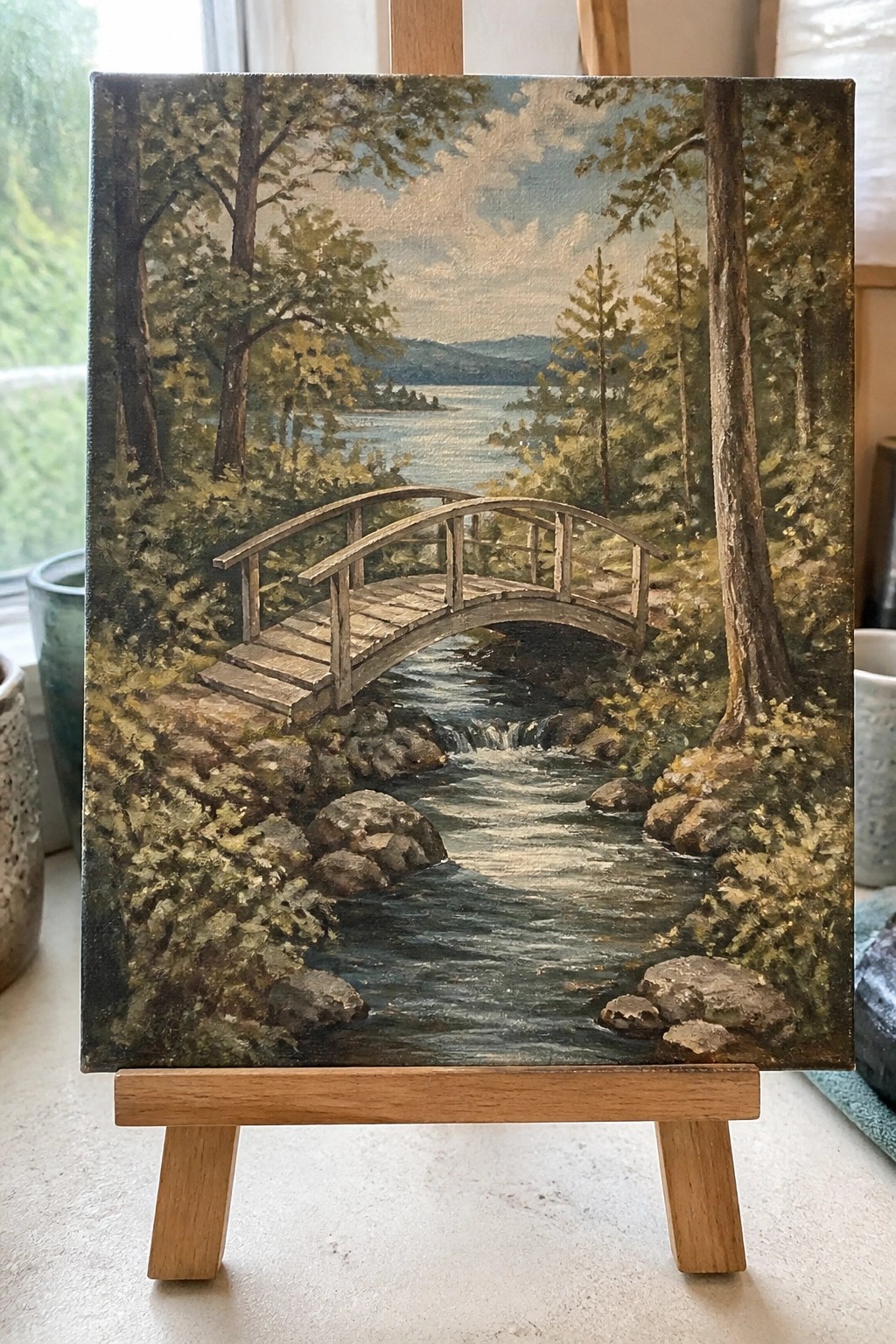 Oil painting on easel showing wooden bridge over rocky stream in forested lakeside scene.
