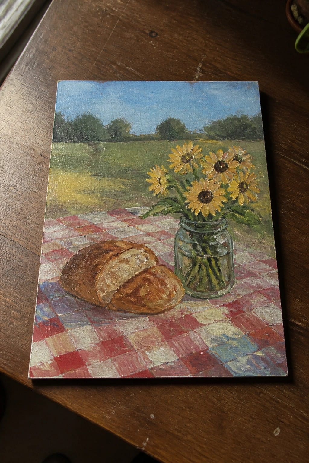 Loaf of bread and sunflowers in jar on red checkered cloth with landscape background