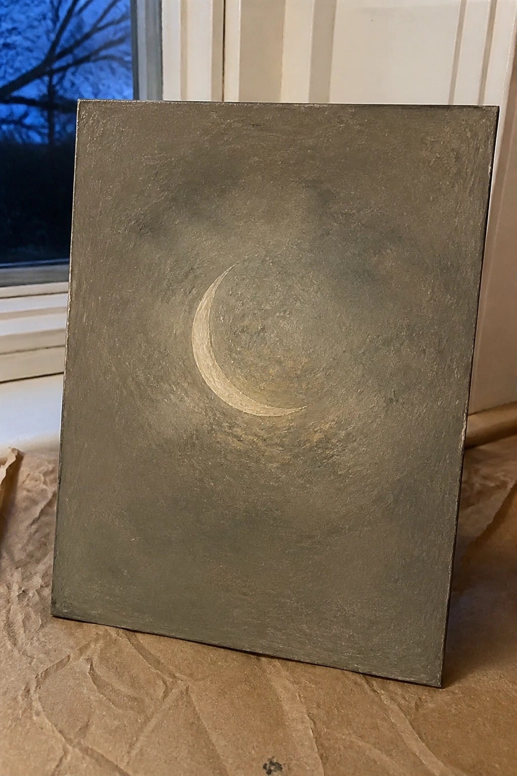 Textured gray canvas painting of a glowing crescent moon propped near a window.