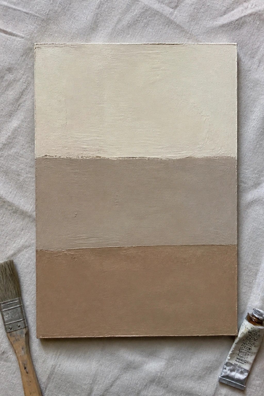 Abstract canvas with three horizontal earth-tone bands beside paintbrush and paint tube on gray fabric