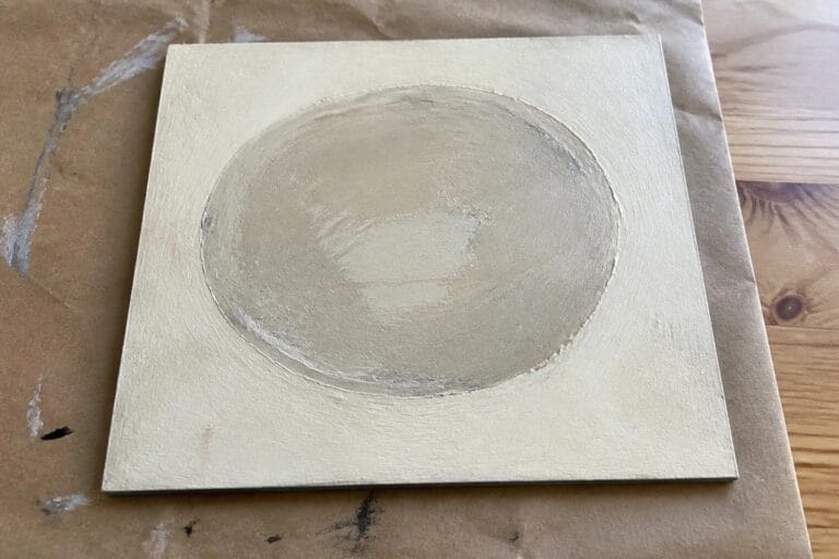 Square canvas with a soft oval painted in light beige tones on brown paper over wood.
