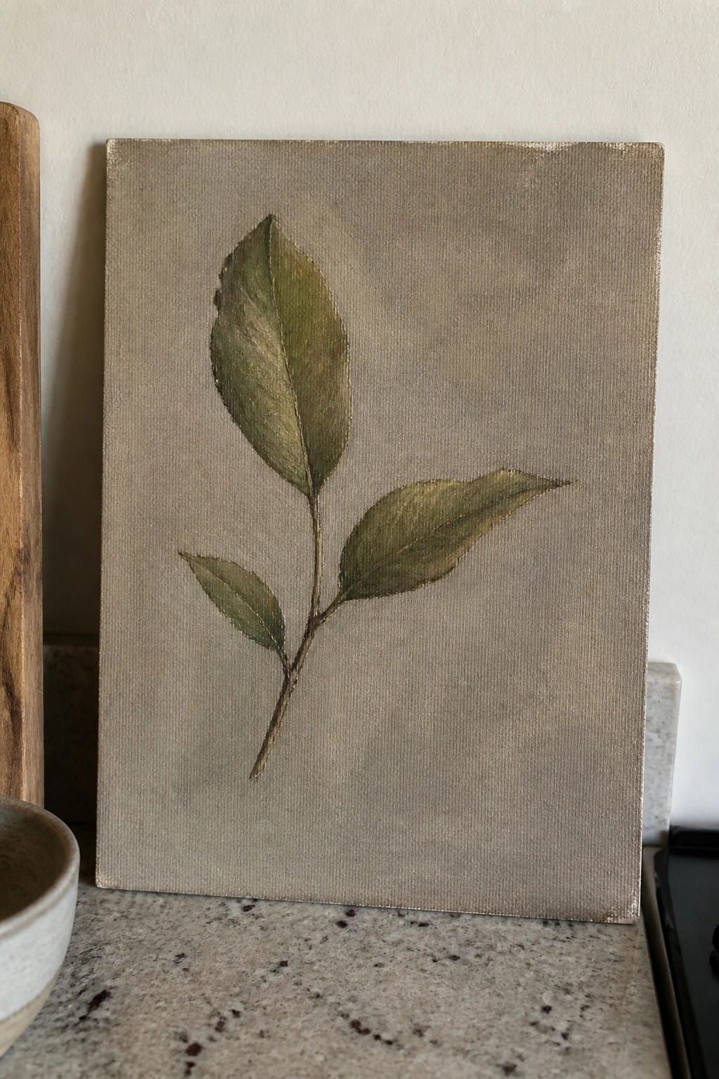 Canvas painting of leafy branch in muted greens on neutral background resting on countertop