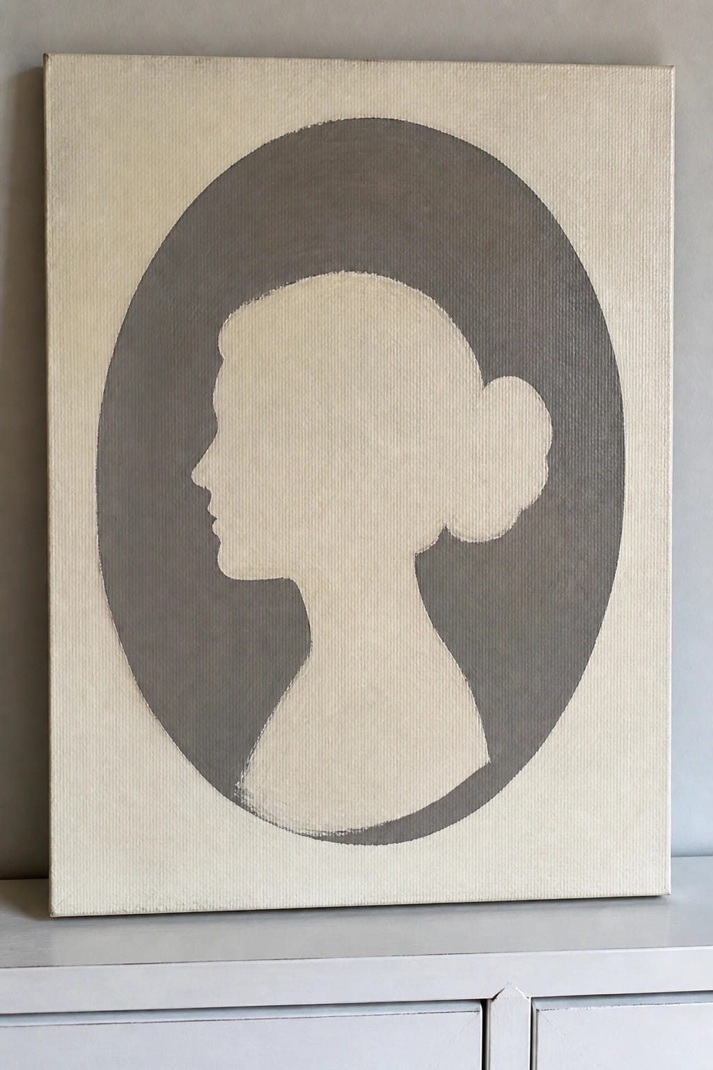 A painting of a woman's profile silhouette in beige inside a gray oval on a light canvas.