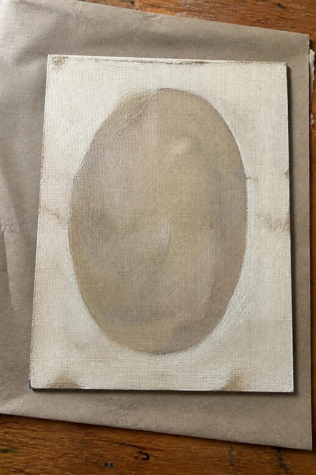 Square canvas with a soft oval painted in light beige tones on brown paper over wood.