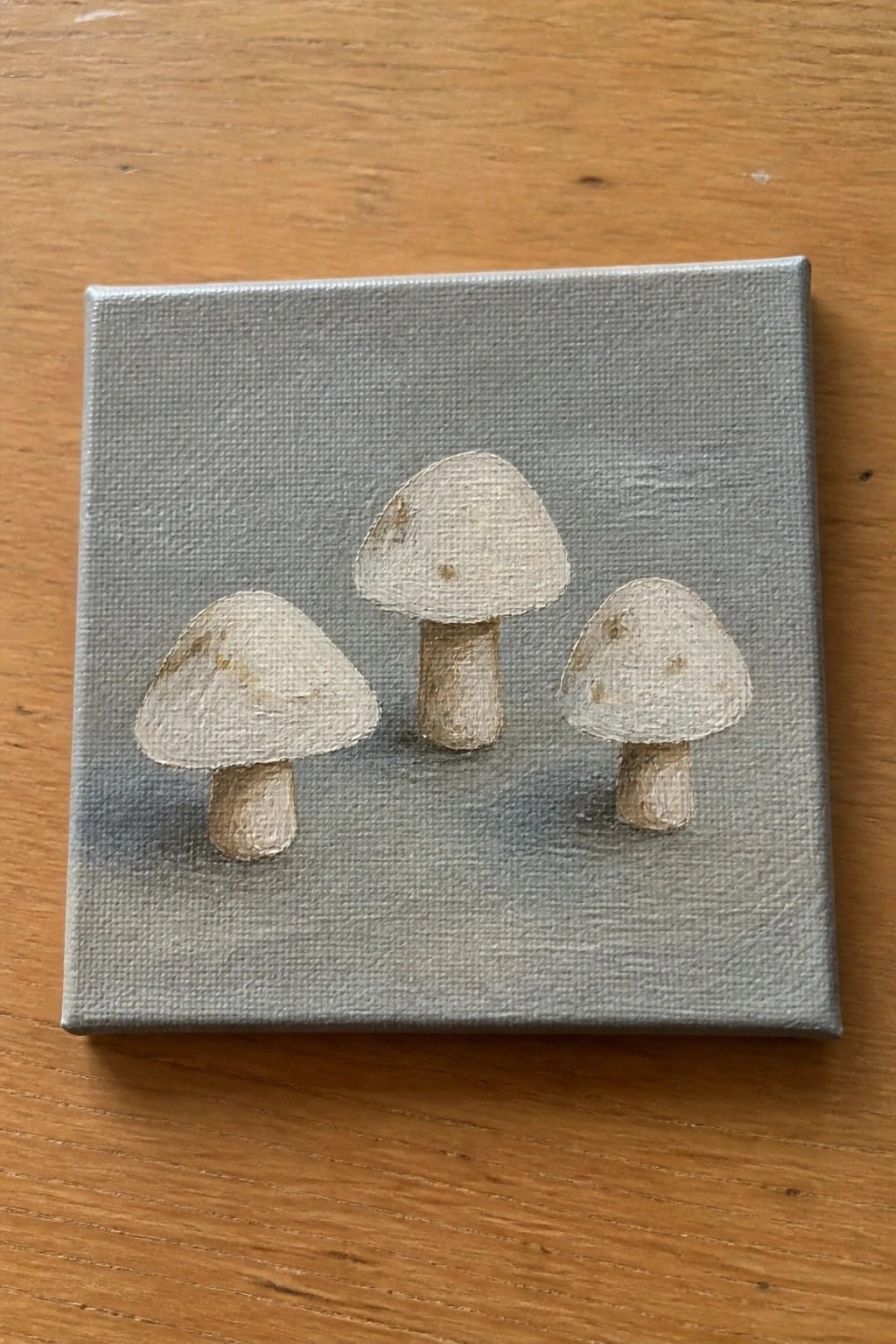 Three pale mushrooms painted on small gray canvas atop wooden table