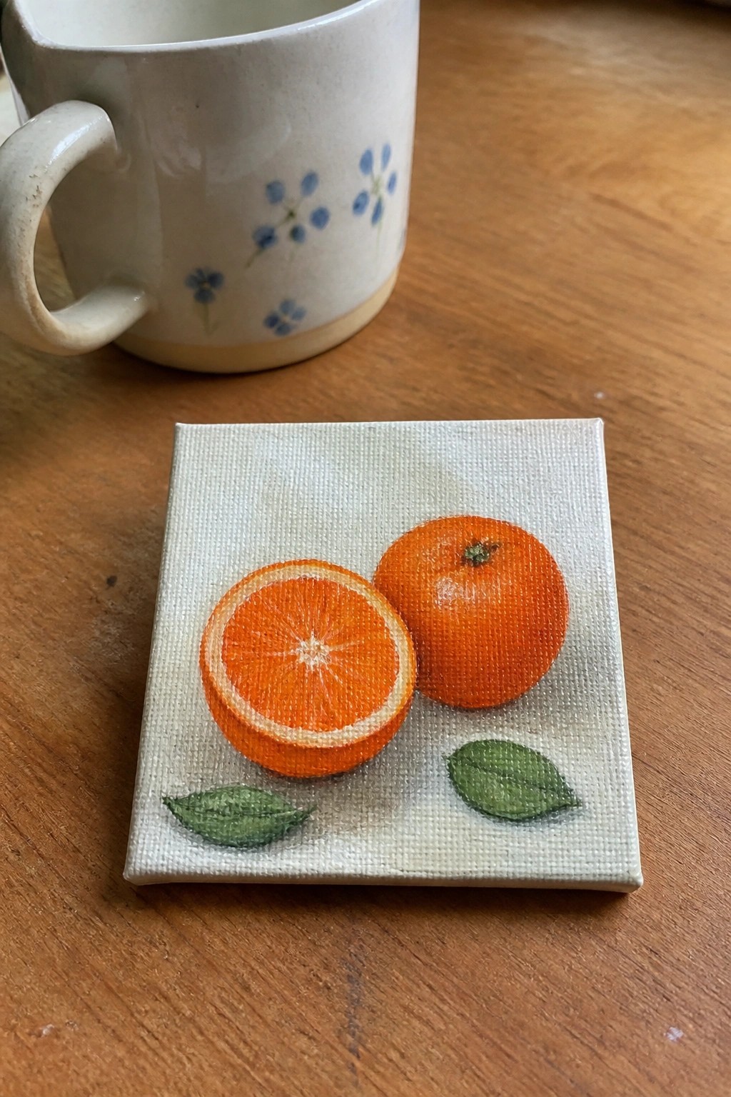 Small canvas painting of whole and halved oranges with leaves beside floral mug on wood
