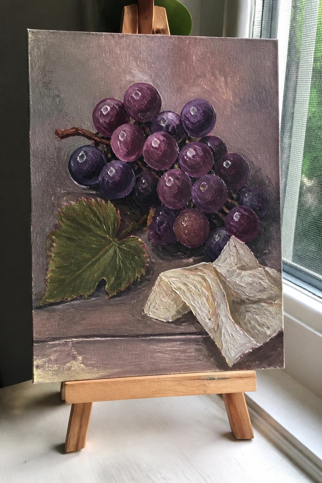 Oil painting of purple grapes with green leaf and white cloth on easel