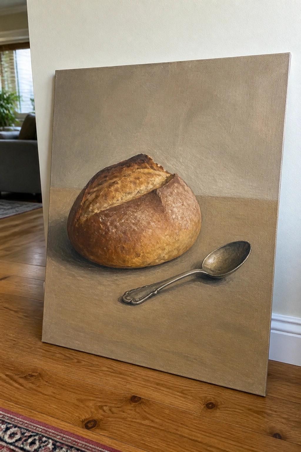 Painting of golden-brown bread loaf and metal spoon on beige canvas in room.