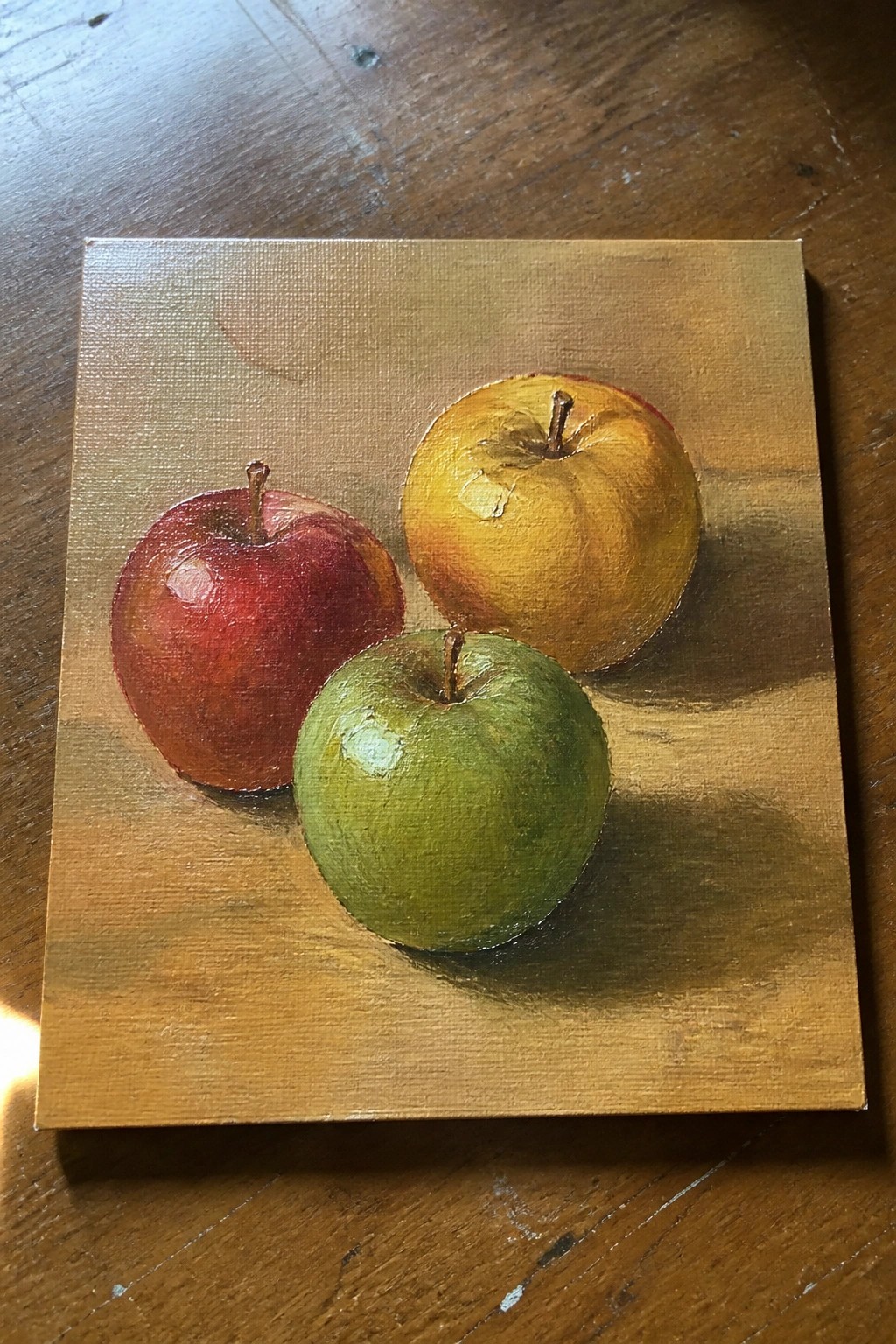 An oil painting of a red apple, a yellow apple, and a green apple on a wooden surface.