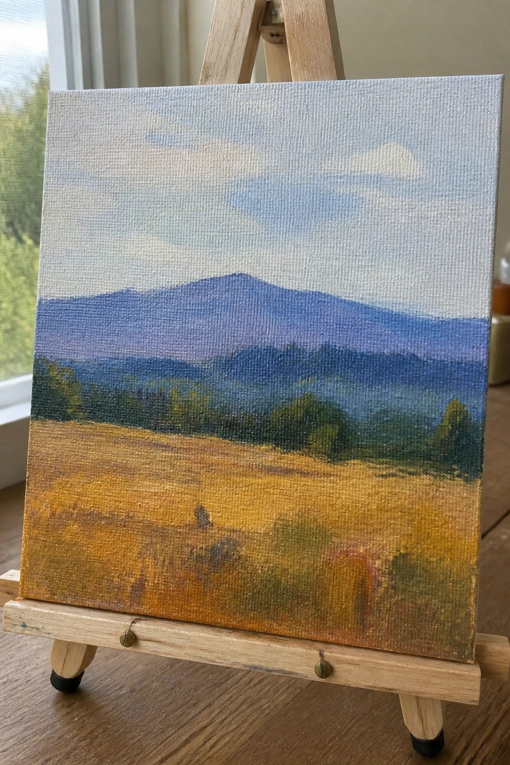 Landscape painting on wooden easel with golden field, green trees, and blue mountains.