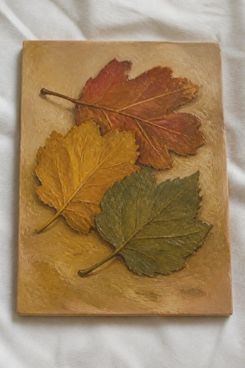Three autumn leaves in red, yellow, and green on a textured brown plaque.