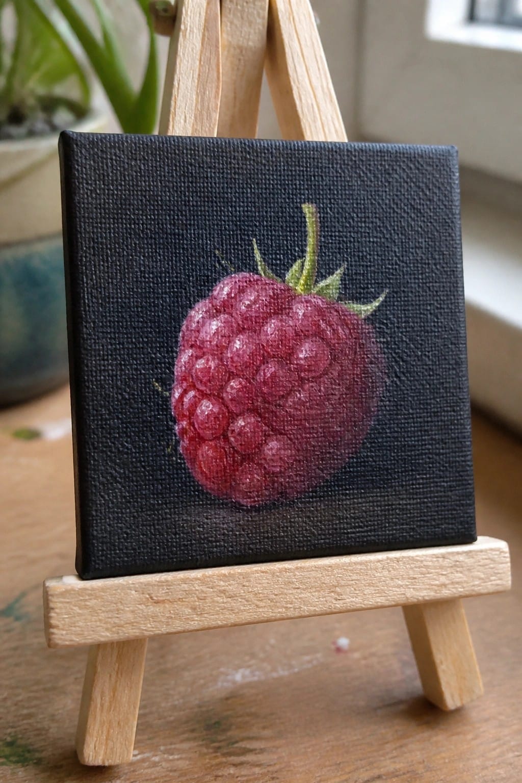 Miniature painting of vibrant red raspberry on black canvas atop wooden easel