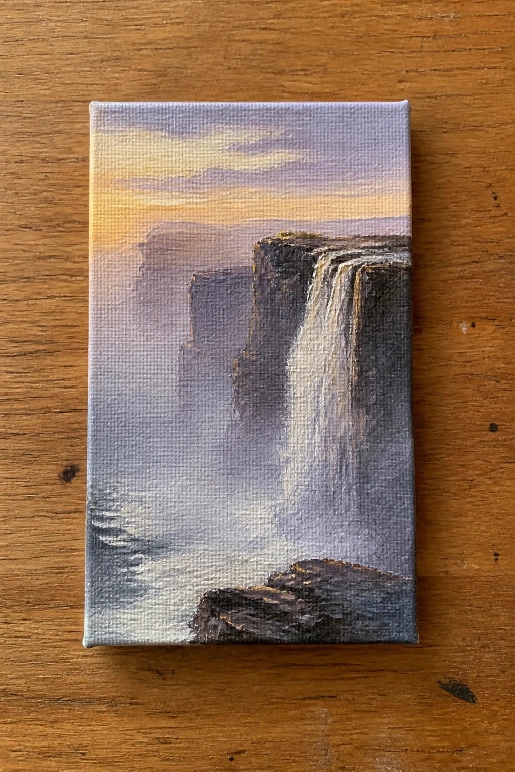 Small canvas painting of misty waterfall cascading from cliffs at sunset on wood.